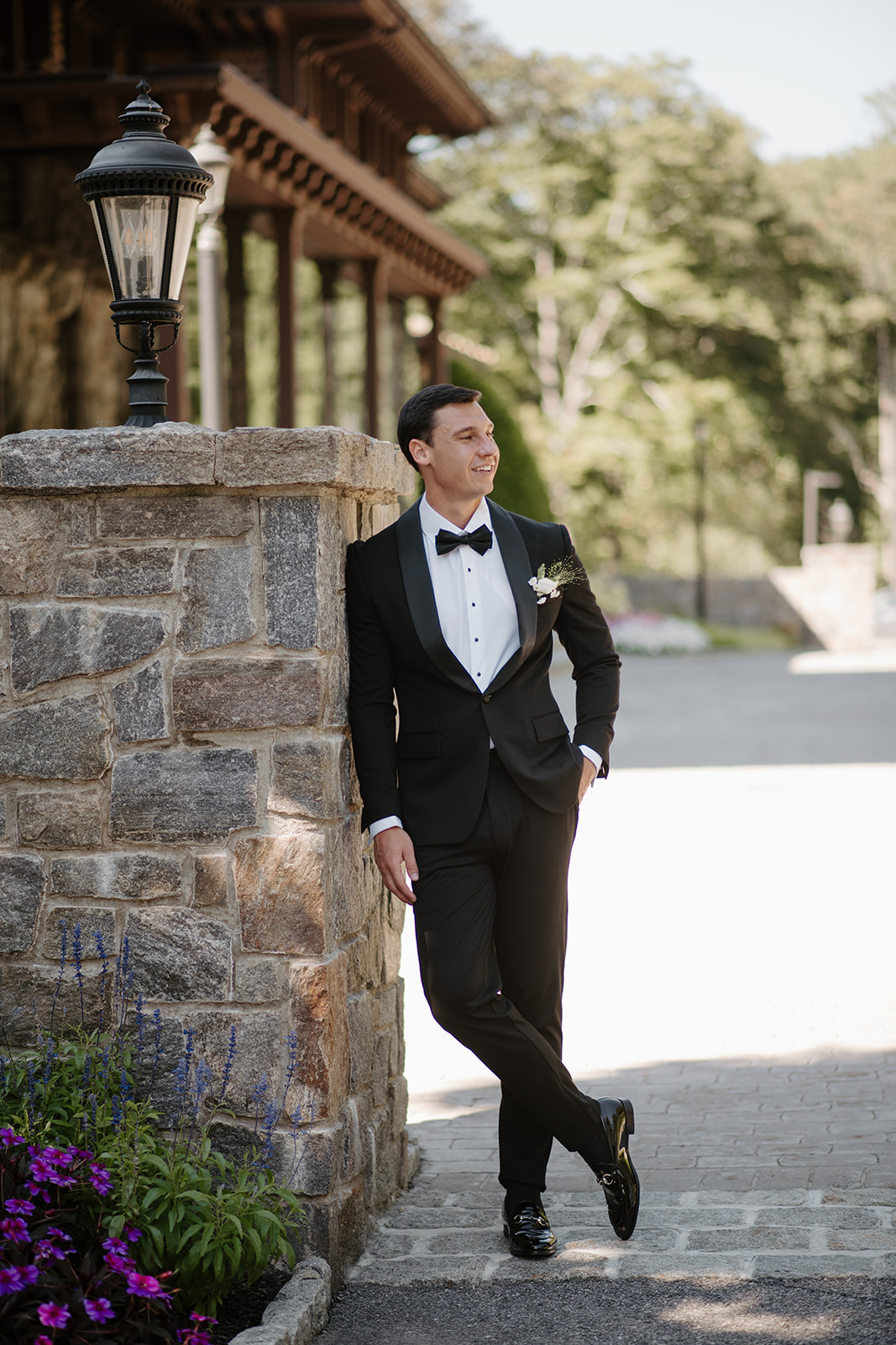 Groom posing confidently while leaning against a stone wall pillar under bright daylight.